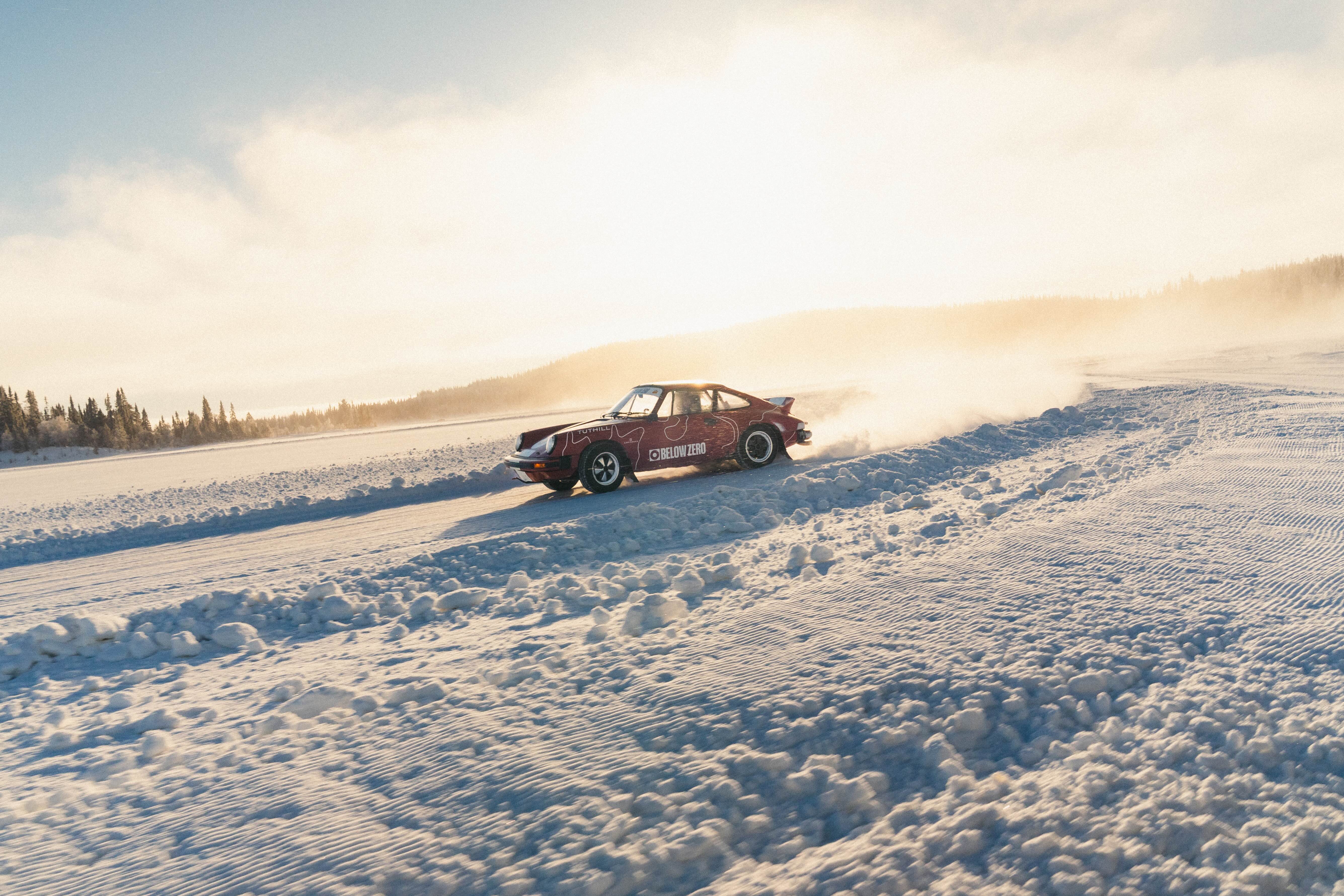 A red Porsche 911 rally car drifts on a snowy track under a bright sky, creating a cloud of snow behind it. Trees are visible in the background.
